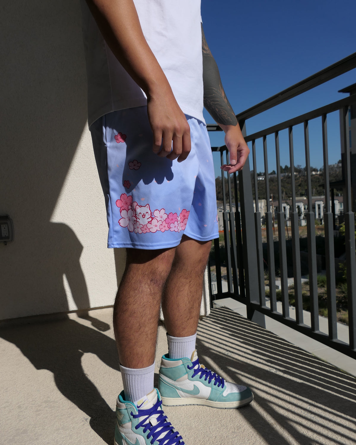 A person standing on a balcony wearing light blue shorts with white and pink cherry blossom an a cute white cat, a white t-shirt, white socks, and mint green and white sneakers with bright blue laces.