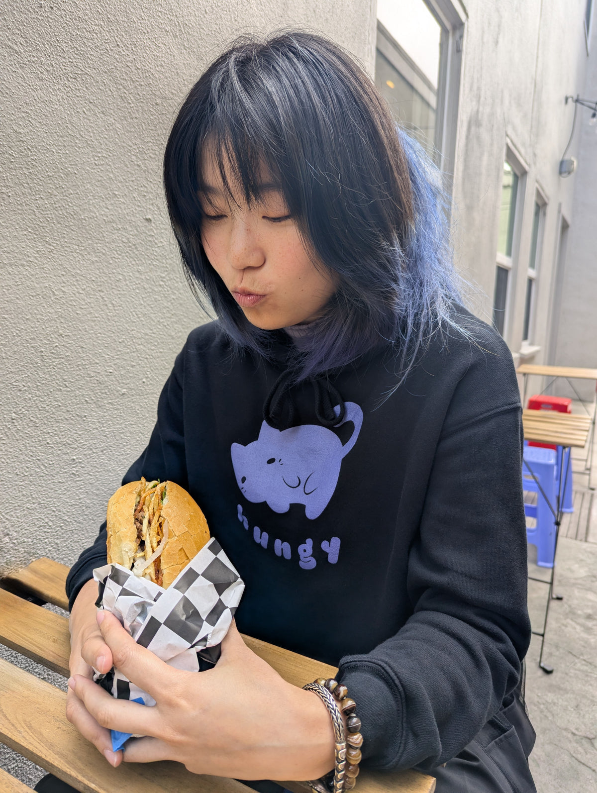 Person wearing a black hoodie with a purple cat graphic design, holding a sandwich outdoors.
